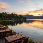 Best Restaurants in Lake Toba: Where to Eat (2025 Guide) A wide shot of a serene Lake Toba at sunset, with traditional Batak houses visible on the shore and a restaurant patio subtly lit, hinting at dining experiences.