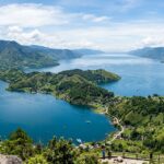 Things to Do in Lake Toba: Top Attractions & Travel Guide 2025 A wide, panoramic view of the vast Lake Toba caldera, with lush green hills surrounding the deep blue waters and Samosir Island visible in the distance under a clear sky.