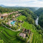 Best Resorts in Ubud, Bali: Top Luxury & Wellness Stays An aerial view of a luxurious resort nestled within lush green rice paddies and tropical jungle in Ubud, Bali, with a dramatic river gorge in the background.