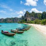 Longtail boats floating in clear turquoise water near a white sand beach with limestone cliffs in Phuket.