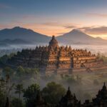 A wide shot of Borobudur Temple at sunrise with mist, surrounded by lush green trees and distant mountains.