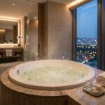 10 Best Hotels With Jacuzzi in Room for 2025 (Suites Ranked) A luxurious modern hotel suite with a large, inviting jacuzzi tub next to a window overlooking a cityscape at dusk.