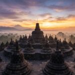 A wide shot of Borobudur temple at sunrise, with mist rising over the surrounding trees.