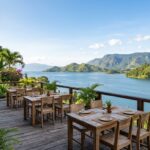 A scenic view of Lake Toba with a restaurant patio by the water's edge, framed by lush hills.